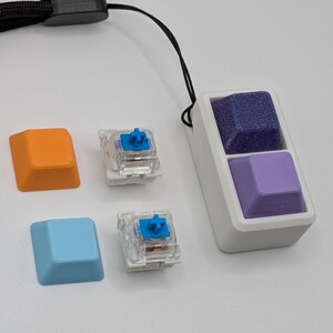 May include: A white rectangular device with two purple keycaps, one textured and one smooth, attached to a black lanyard. Two mechanical keyboard switches and two additional keycaps in orange and blue are also present.