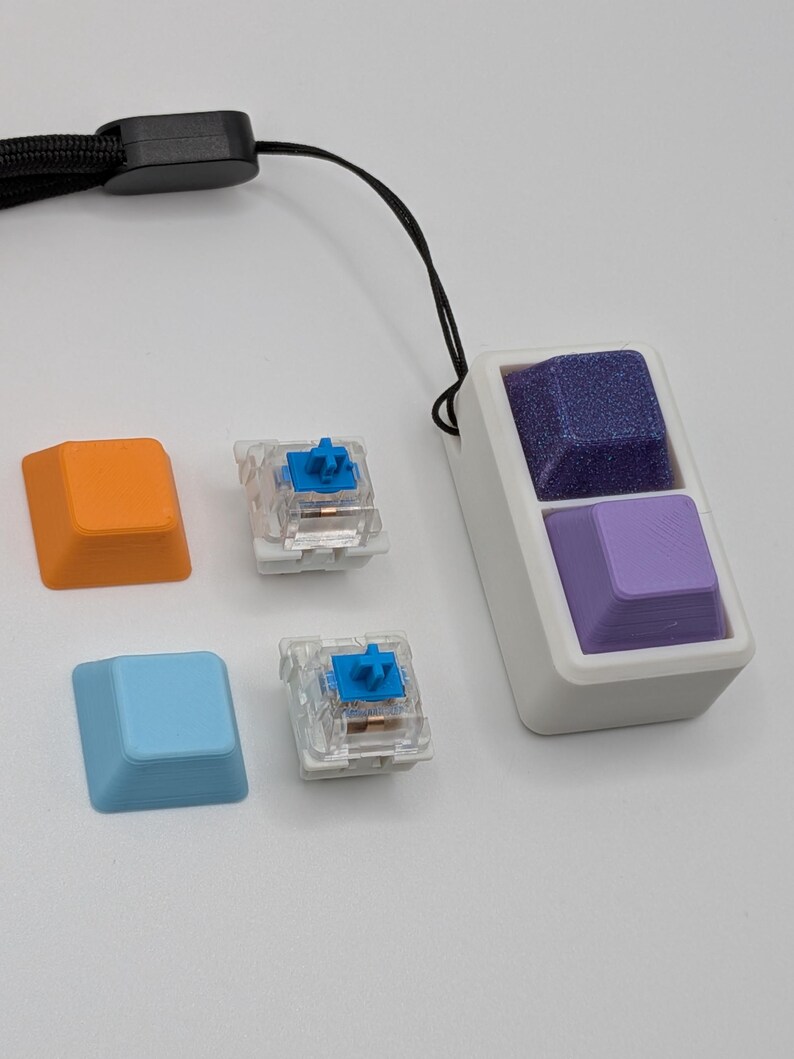 May include: A white rectangular device with two purple keycaps, one textured and one smooth, attached to a black lanyard. Two mechanical keyboard switches and two additional keycaps in orange and blue are also present.