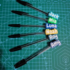 May include: Six black ballpoint pens with personalized name tags in various colors. The names include Jack, Mia, Luna, Noah, Roy, and Amy. The pens are arranged on a green cutting mat with measurement markings.