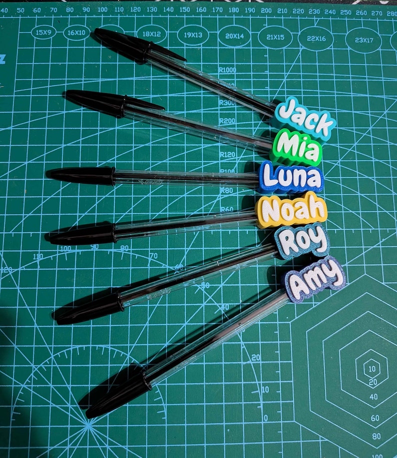 May include: Six black ballpoint pens with personalized name tags in various colors. The names include Jack, Mia, Luna, Noah, Roy, and Amy. The pens are arranged on a green cutting mat with measurement markings.