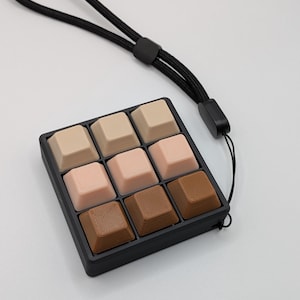 May include: A nine-key keyboard keycap set in a black tray with a black wrist strap. The keycaps are square and in shades of beige, pink, and brown. The wrist strap is made of a black woven material.