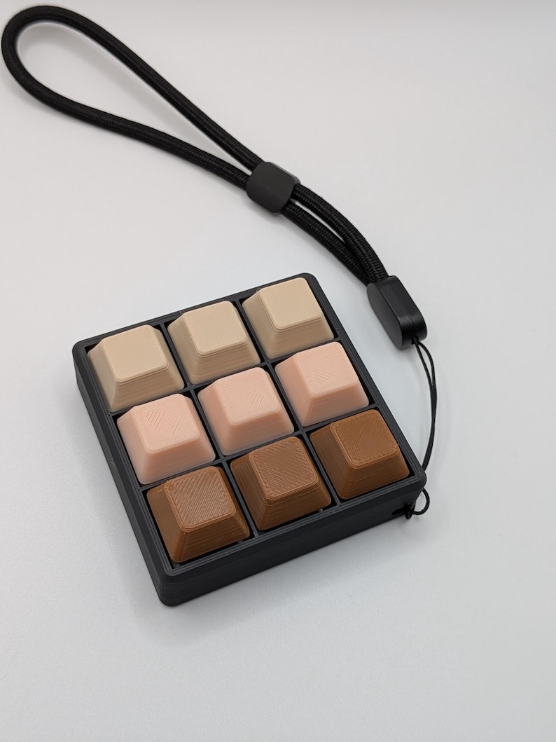 May include: A nine-key keyboard keycap set in a black tray with a black wrist strap. The keycaps are square and in shades of beige, pink, and brown. The wrist strap is made of a black woven material.