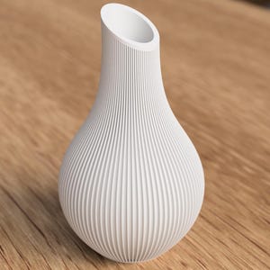 May include: A white, textured vase with a bulbous base and a flared, angled opening. The vase has vertical, ridged detailing, creating a ribbed effect. The vase is set against a wooden surface.