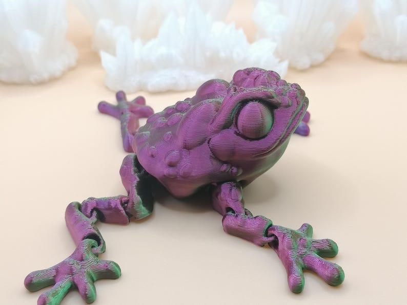May include: A 3D-printed articulated frog toy in shades of purple and green. The frog has a textured surface and detailed features, including large eyes and jointed limbs. The background features white, crystalline formations.