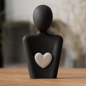 Puede incluir: Escultura negra de una persona sosteniendo un coraz&oacute;n blanco con l&iacute;neas verticales. La escultura tiene una superficie texturizada y est&aacute; sobre una mesa de madera. El fondo est&aacute; borroso, con toques de vegetaci&oacute;n y tonos neutros.