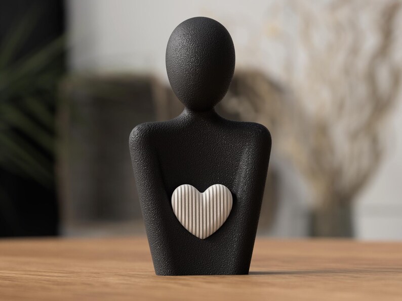 Puede incluir: Escultura negra de una persona sosteniendo un coraz&oacute;n blanco con l&iacute;neas verticales. La escultura tiene una superficie texturizada y est&aacute; sobre una mesa de madera. El fondo est&aacute; borroso, con toques de vegetaci&oacute;n y tonos neutros.