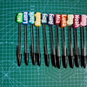 May include: A collection of ten black ballpoint pens, each topped with a colorful name tag. The name tags feature names like Mia, Luna, Jack, and Noah in various colors. The pens are arranged on a green cutting mat.
