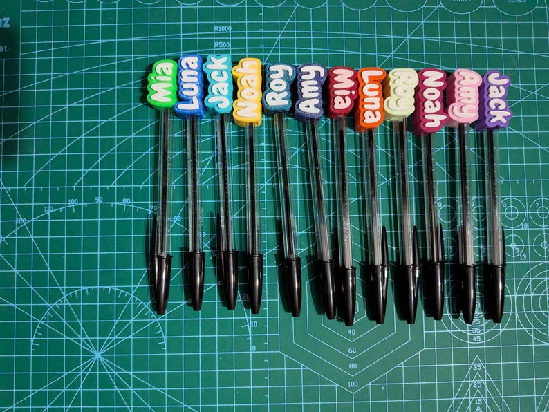 May include: A collection of ten black ballpoint pens, each topped with a colorful name tag. The name tags feature names like Mia, Luna, Jack, and Noah in various colors. The pens are arranged on a green cutting mat.
