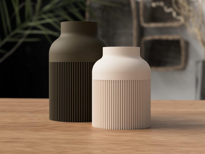 May include: Two modern ceramic vases. One is olive green, the other is cream. Both have a cylindrical base with vertical ribbed detailing. The vases sit on a wooden surface, with a blurred background of greenery and neutral tones.