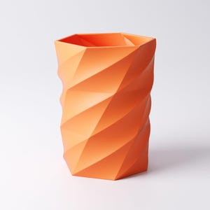 May include: An orange geometric vase with a faceted design. The vase has a cylindrical shape with a hexagonal top and a repeating pattern of angled planes. The color is a vibrant, solid orange.