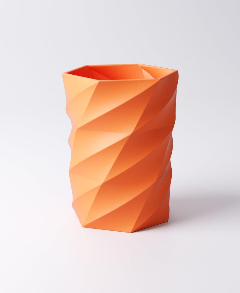 May include: An orange geometric vase with a faceted design. The vase has a cylindrical shape with a hexagonal top and a repeating pattern of angled planes. The color is a vibrant, solid orange.