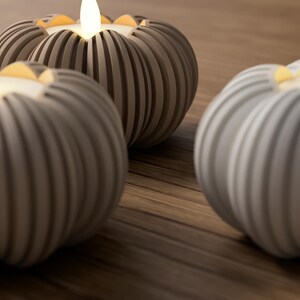 May include: Three gray, ribbed, pumpkin-shaped candle holders with lit, warm-toned LED candles. The candle holders are arranged on a wooden surface, creating a cozy and decorative ambiance. The soft glow of the candles adds a touch of warmth.