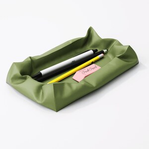 May include: An olive green desk tray with a wavy design, holding a black and white pen, a yellow pencil, and a pink eraser with the text "Pink Pearl". The tray is rectangular and sits on a white surface.