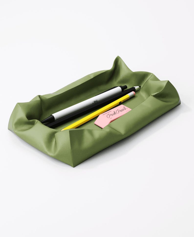 May include: An olive green desk tray with a wavy design, holding a black and white pen, a yellow pencil, and a pink eraser with the text "Pink Pearl". The tray is rectangular and sits on a white surface.