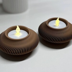 May include: Two brown, decorative candle holders with a swirling design. Each holder contains a white candle with a flickering yellow LED flame. The holders are on a white surface, suggesting a home decor item.