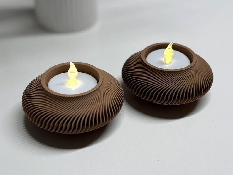 May include: Two brown, decorative candle holders with a swirling design. Each holder contains a white candle with a flickering yellow LED flame. The holders are on a white surface, suggesting a home decor item.