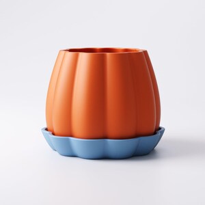 May include: Orange ceramic pumpkin-shaped planter with vertical ridges, sitting on a blue scalloped saucer. The planter has a wide opening and a smooth, glossy finish. A decorative item for home decor.