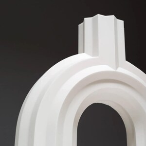 May include: A white, abstract sculpture with a rounded arch and a rectangular top. The sculpture has a minimalist design with smooth, curved lines and a matte finish. The background is a dark gray.