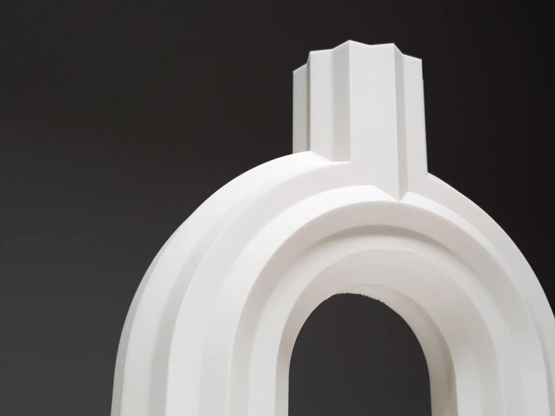 May include: A white, abstract sculpture with a rounded arch and a rectangular top. The sculpture has a minimalist design with smooth, curved lines and a matte finish. The background is a dark gray.