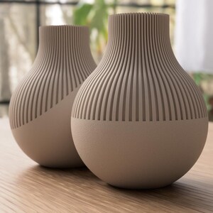 May include: Two beige ceramic vases with a ribbed design on the upper portion. The vases have a rounded shape and sit on a wooden surface. The background is blurred, suggesting a natural setting.