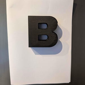 May include: A black, three-dimensional letter "B" with a textured surface. The letter is positioned on a white sheet of paper, casting a shadow. The background is a dark gray surface.