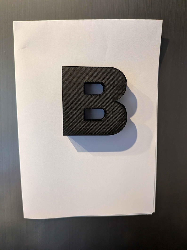 May include: A black, three-dimensional letter "B" with a textured surface. The letter is positioned on a white sheet of paper, casting a shadow. The background is a dark gray surface.