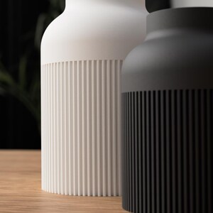 May include: Two modern ceramic vases. One is white, the other is black. Both have a cylindrical ribbed design at the base. The vases are on a wooden surface, with a blurred background.