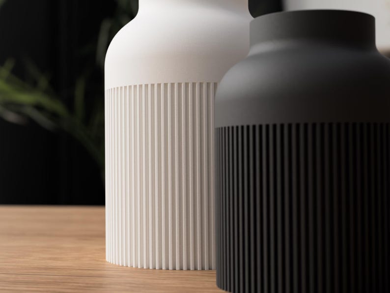 May include: Two modern ceramic vases. One is white, the other is black. Both have a cylindrical ribbed design at the base. The vases are on a wooden surface, with a blurred background.