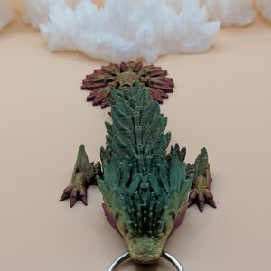 May include: A detailed, 3D-printed dragon head pendant in shades of green, gold, and burgundy. The dragon has a textured, scaled appearance with a silver ring attached at the bottom. The background features white crystal formations and a light beige surface.