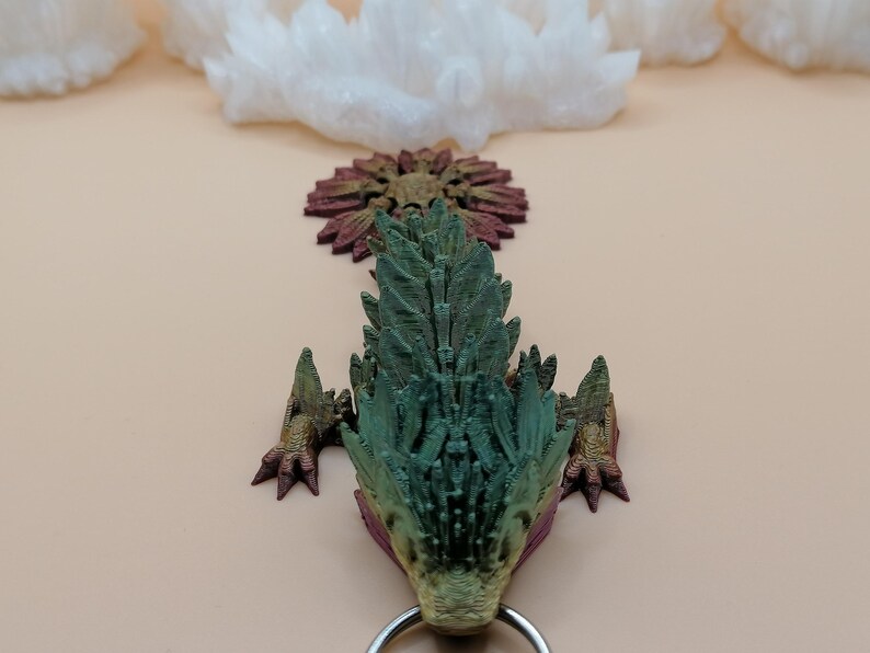 May include: A detailed, 3D-printed dragon head pendant in shades of green, gold, and burgundy. The dragon has a textured, scaled appearance with a silver ring attached at the bottom. The background features white crystal formations and a light beige surface.