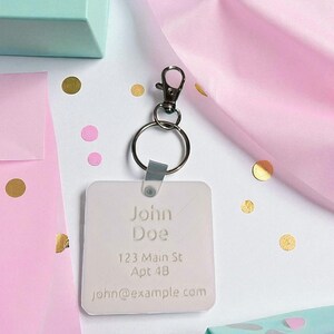 May include: A personalized keychain with the name "John Doe", address, and email. The keychain is square-shaped and attached to a silver-colored clasp and ring. The background includes gift boxes, confetti, and pink tissue paper.