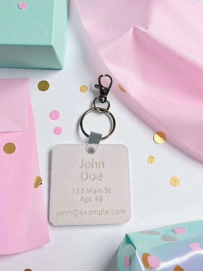 May include: A personalized keychain with the name "John Doe", address, and email. The keychain is square-shaped and attached to a silver-colored clasp and ring. The background includes gift boxes, confetti, and pink tissue paper.