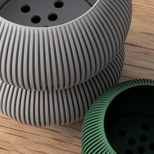 May include: Three modern, ribbed planters in shades of gray and green. The gray planters are stacked, showcasing a detailed, grooved design. The green planter is positioned to the side, offering a contrasting color and texture. The planters sit on a wooden surface.