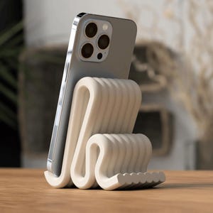 Puede incluir: Un soporte para tel&eacute;fono de color crema con un dise&ntilde;o ondulado, sosteniendo un smartphone plateado. El soporte tiene una superficie texturizada y acanalada, colocado sobre una mesa de madera. El tel&eacute;fono tiene m&uacute;ltiples lentes.
