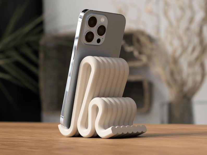 Puede incluir: Un soporte para tel&eacute;fono de color crema con un dise&ntilde;o ondulado, sosteniendo un smartphone plateado. El soporte tiene una superficie texturizada y acanalada, colocado sobre una mesa de madera. El tel&eacute;fono tiene m&uacute;ltiples lentes.