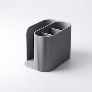 May include: A gray desk organizer with a vertical ribbed texture. The organizer has three compartments and a curved back, designed to hold pens, pencils, or other office supplies. The base is a solid, rounded shape.