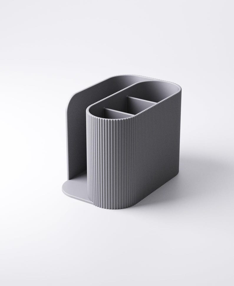 May include: A gray desk organizer with a vertical ribbed texture. The organizer has three compartments and a curved back, designed to hold pens, pencils, or other office supplies. The base is a solid, rounded shape.