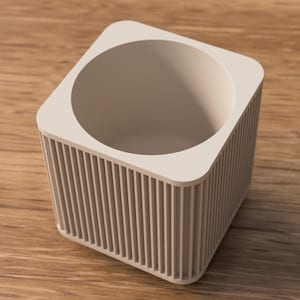 May include: A beige, square planter with a circular opening. The exterior has vertical, ridged detailing. The planter is made of a solid material and sits on a wooden surface.