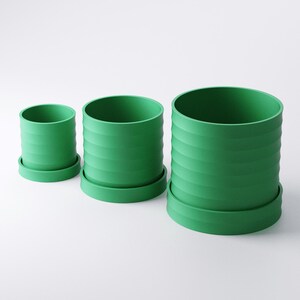 Op de afbeelding: Drie groene cilindrische bloempotten van verschillende formaten, elk met een bijpassende schotel. De potten hebben een geribbeld ontwerp aan de buitenkant. De kleinste pot is ongeveer 10 cm hoog, de middelste pot 15 cm en de grootste 20 cm.