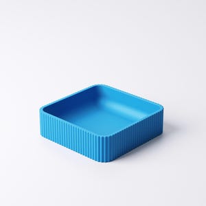 May include: A bright blue, square container with a ribbed exterior. The container has a simple, modern design and is likely made of plastic or a similar material. The interior is smooth and empty.