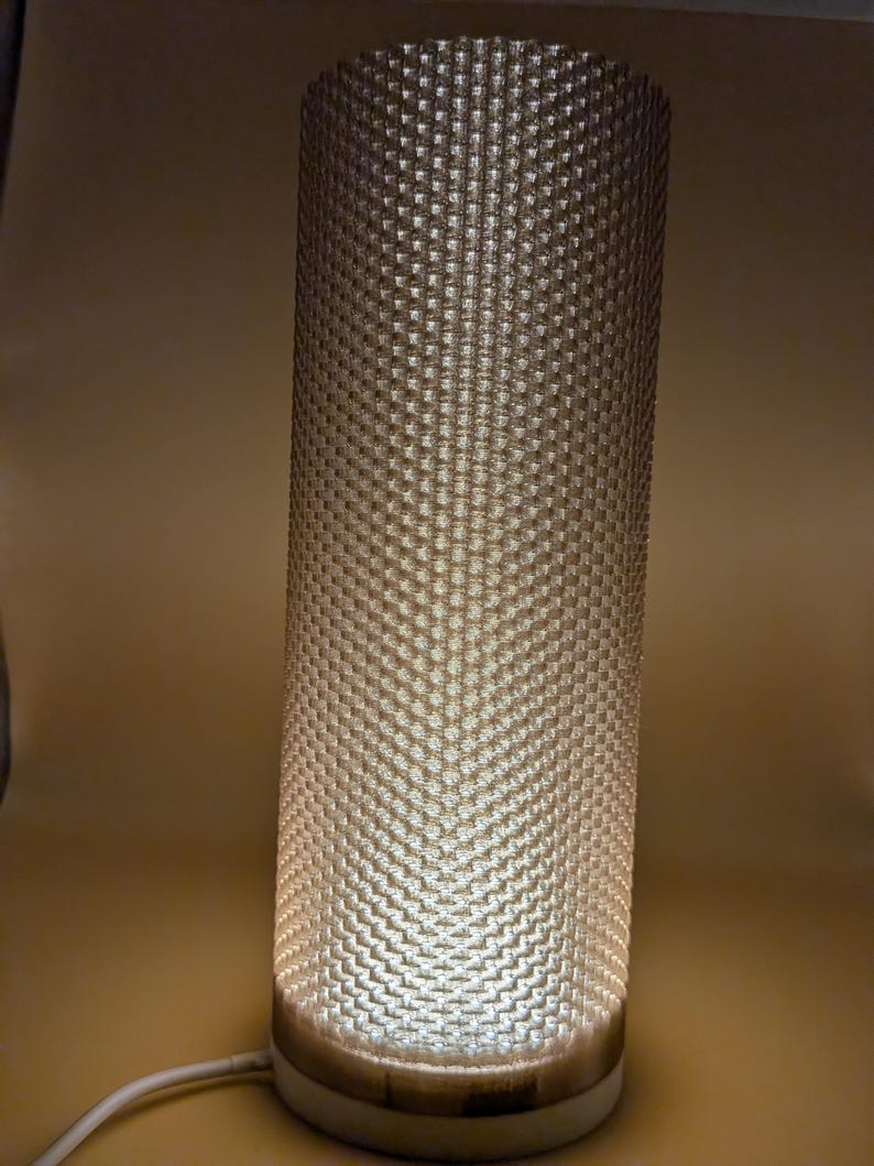 K&ouml;nnte beinhalten: Zylindrische Lampe mit strukturiertem, gemustertem Schirm. Der Schirm hat ein sich wiederholendes quadratisches Muster und wird von innen beleuchtet, wodurch ein sanftes Leuchten entsteht. Der Sockel ist hell und ein wei&szlig;es Kabel geht vom Boden aus.