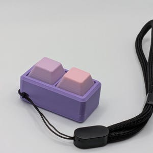 May include: A purple rectangular case holds two gradient-colored keycaps, one pink and one lavender. A black lanyard with a black plastic clasp is attached to the case. The keycaps are designed for a mechanical keyboard.