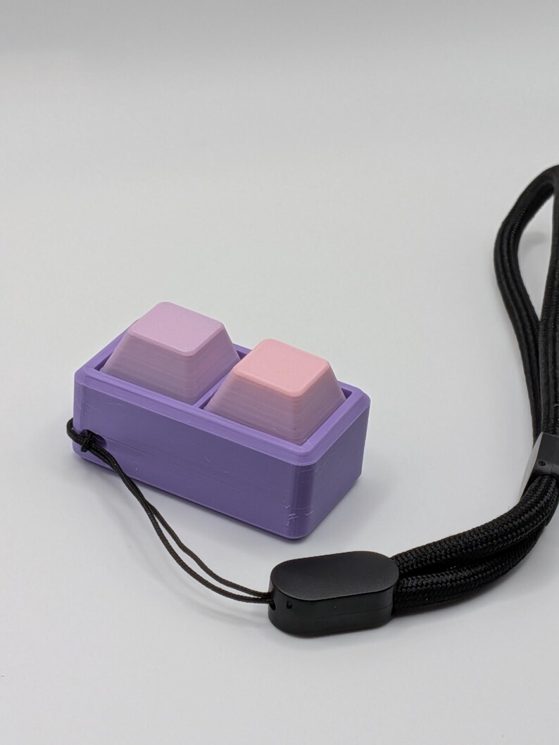 May include: A purple rectangular case holds two gradient-colored keycaps, one pink and one lavender. A black lanyard with a black plastic clasp is attached to the case. The keycaps are designed for a mechanical keyboard.