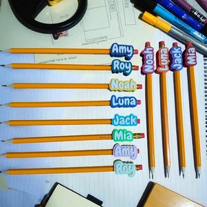 May include: A collection of yellow pencils with attached name tags. The name tags are in various colors and feature names like Amy, Roy, Noah, Luna, Jack, and Mia. The pencils are arranged on a white surface with other stationery items.