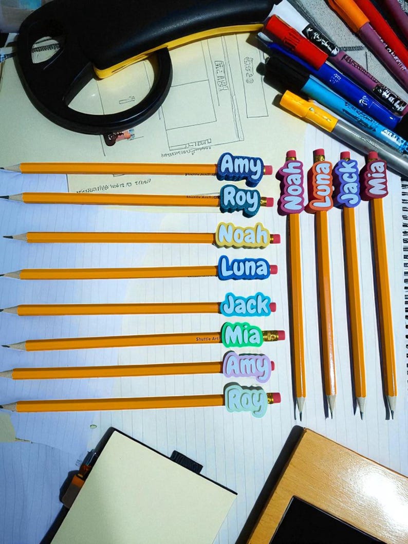 May include: A collection of yellow pencils with attached name tags. The name tags are in various colors and feature names like Amy, Roy, Noah, Luna, Jack, and Mia. The pencils are arranged on a white surface with other stationery items.