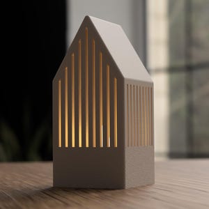 May include: A small, house-shaped lamp in a neutral gray color. The lamp has vertical slits that allow warm light to shine through. It is sitting on a wooden surface, with a blurred background.