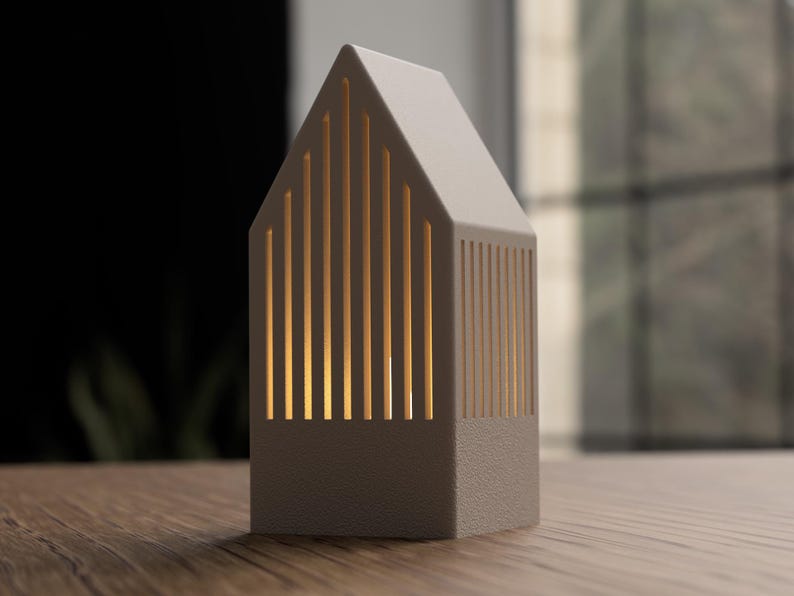 May include: A small, house-shaped lamp in a neutral gray color. The lamp has vertical slits that allow warm light to shine through. It is sitting on a wooden surface, with a blurred background.
