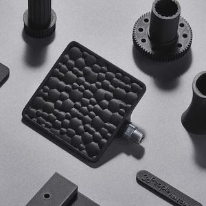 May include: An assortment of black 3D-printed mechanical parts, including a textured square pedal, a gear, a multi-holed cylinder, and other geometric shapes. The parts are arranged on a light gray surface.