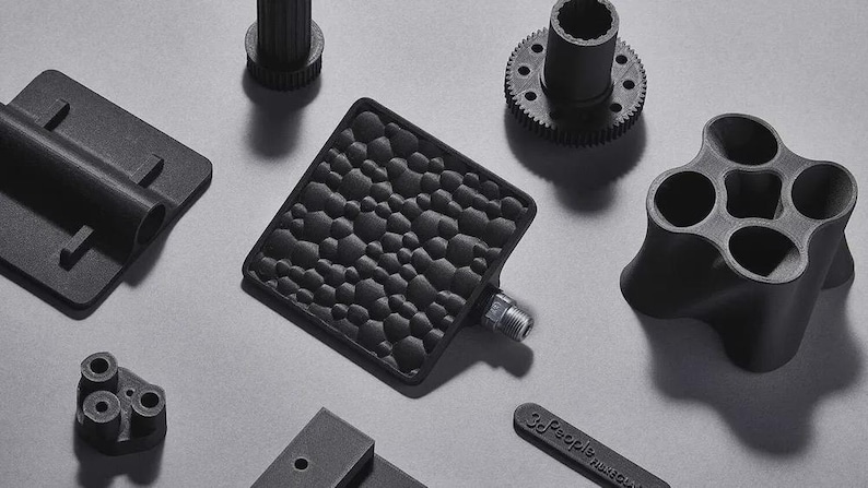 May include: An assortment of black 3D-printed mechanical parts, including a textured square pedal, a gear, a multi-holed cylinder, and other geometric shapes. The parts are arranged on a light gray surface.