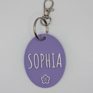May include: A lavender oval keychain with the name "SOPHIA" in white, accompanied by a small flower graphic. The keychain has a silver-colored clasp and ring for attachment.